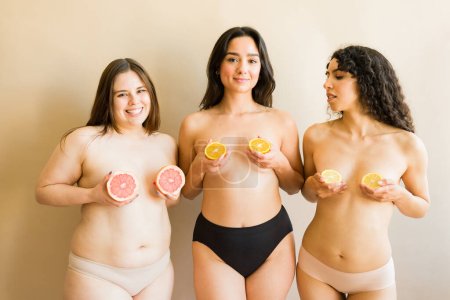 Téléchargez les photos : Bonnes femmes heureuses avec leurs poitrines nues couvrant leur poitrine d'oranges et de pamplemousses célébrant la positivité du corps - en image libre de droit