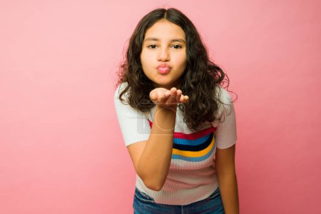Téléchargez les photos : Belle hispanique adolescent fille souffler un baiser et avoir du plaisir à la recherche heureux sur un fond rose - en image libre de droit