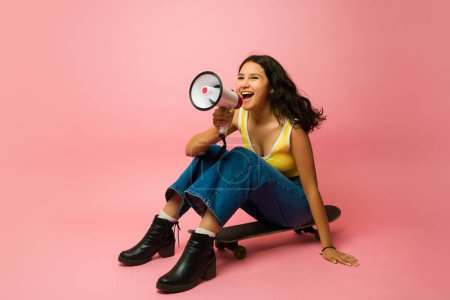 Foto de Emocionada chica de trece años gritando un mensaje con un megáfono mirando alegre mientras se sienta en un monopatín - Imagen libre de derechos