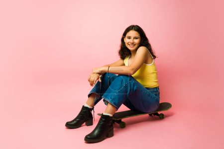 Photo for Cheerful happy teen girl ready to start skating and playing with a skateboard while smiling against a pink background - Royalty Free Image