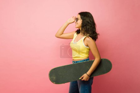 Foto de Vista lateral de una adolescente latina feliz poniéndose gafas de sol retro y llevando un monopatín vintage listo para empezar a patinar - Imagen libre de derechos