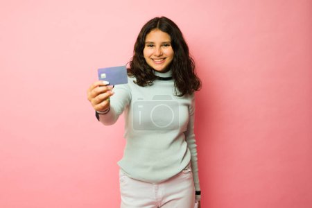 Foto de Emocionada adolescente hispana mostrando su nueva primera tarjeta de crédito y pagando mientras va de compras con un fondo rosa - Imagen libre de derechos