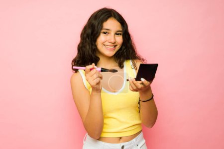 Téléchargez les photos : Heureuse adolescente latine contact visuel tout en souriant et en se maquillant se préparant à sortir - en image libre de droit