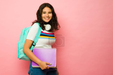 Téléchargez les photos : Heureux adolescent de treize ans portant un sac à dos et des livres tout en souriant aller au collège à côté de l'espace de copie rose - en image libre de droit