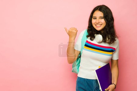 Téléchargez les photos : Étudiante adolescente intelligente portant ses livres scolaires et son sac à dos pointant vers une annonce d'espace de copie rose et souriant - en image libre de droit