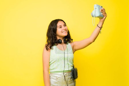 Téléchargez les photos : Sourire adolescent fille prendre un selfie à l'aide d'un appareil photo instantané et avoir du plaisir en été sur un fond jaune - en image libre de droit