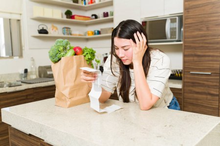 Photo for Worried woman in the kitchen feeling stressed looking at the expensive receipt after buying food at the supermarket during bad economy - Royalty Free Image