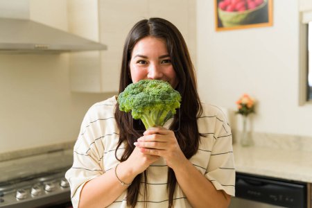 Foto de Atractiva mujer vegana haciendo contacto visual y sonriendo mientras muestra un brócoli vegetal verde listo para cocinar alimentos saludables - Imagen libre de derechos