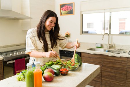 Foto de Relajada mujer vegetariana sonriente en la cocina preparando una receta saludable con comida vegana y verduras orgánicas - Imagen libre de derechos