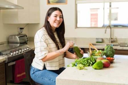 Téléchargez les photos : Joyeux belle femme manger une salade verte saine avec des légumes biologiques et boire du jus vert dans la cuisine - en image libre de droit