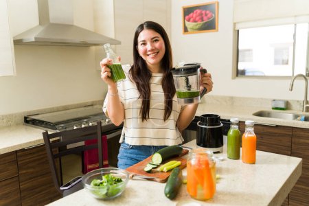 Téléchargez les photos : Magnifique femme en bonne santé souriant tout en buvant un jus vert ou un smoothie avec des légumes biologiques dans sa cuisine - en image libre de droit
