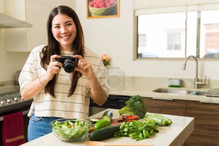 Photo for Happy vegan woman taking photos with a camera while cooking healthy vegetarian food in the kitchen - Royalty Free Image