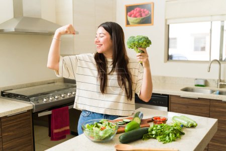 Photo for Healthy vegetarian woman feeling strong while eating organic vegetables and cooking a vegan recipe in the kitchen - Royalty Free Image