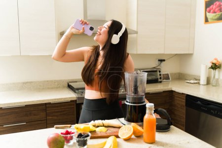 Foto de Mujer vegana alegre cantando escuchando música con auriculares y divirtiéndose mientras bebe un batido de frutas orgánicas - Imagen libre de derechos