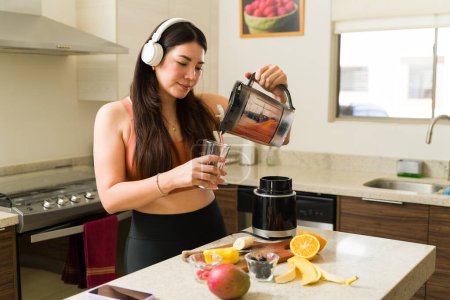Foto de Fit mujer vegana activa sonriendo y bebiendo un batido de frutas orgánicas en la cocina después de hacer ejercicio - Imagen libre de derechos