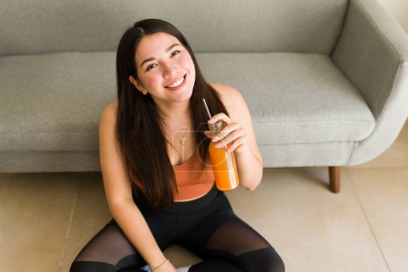 Photo for High angle of a beautiful caucasian woman drinking a healthy carrot juice after a fitness workout at home - Royalty Free Image