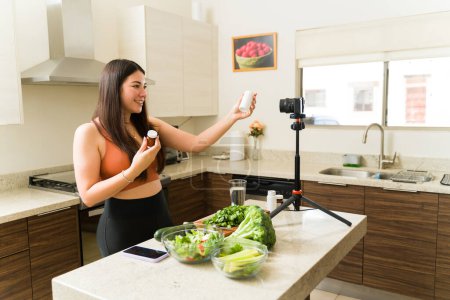 Téléchargez les photos : Fitness jeune femme filmant une vidéo tout en cuisinant avec des légumes biologiques et montrant ses pilules de vitamines - en image libre de droit