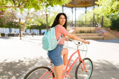 Foto de Mujer hispana feliz con una mochila con pantalones cortos sonriendo vista desde atrás mientras monta su bicicleta en el parque durante el verano - Imagen libre de derechos