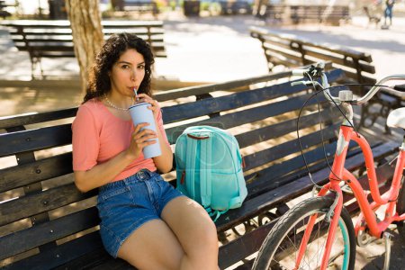 Photo for Latin attractive young woman drinking coffee or water while sitting on the park bench with her bicycle and backpack in the summer - Royalty Free Image