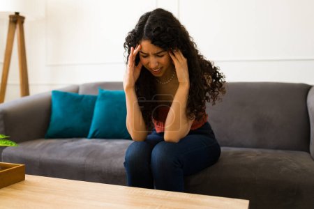 Foto de Mujer joven hispana estresada frotando sus sienes y sufriendo de un dolor de cabeza o migraña en la sala de estar - Imagen libre de derechos