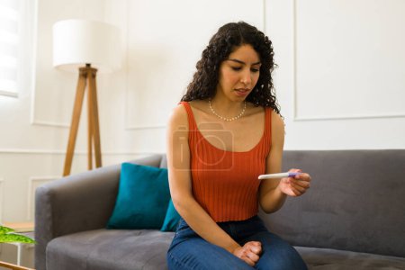 Téléchargez les photos : Jeune femme inquiète regardant triste et stressé en regardant les résultats du test de grossesse dans sa maison - en image libre de droit