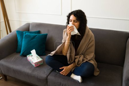 Foto de Mujer joven hispana enferma sonándose la nariz y enferma con un resfriado o gripe envuelta en una manta en casa - Imagen libre de derechos