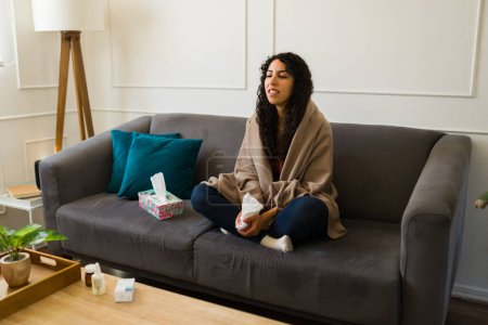 Foto de Mujer latina cansada de 20 años envuelta en una manta y enferma con un resfriado o la gripe sintiéndose mal y mal en el sofá - Imagen libre de derechos