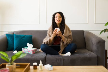Téléchargez les photos : Jeune femme malade énervée utilisant des tissus lui mouchant le nez enveloppé dans une couverture tout en se sentant malade d'un mauvais rhume ou de la grippe - en image libre de droit
