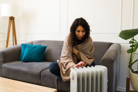 Foto de Mujer joven incómoda sintiéndose fría envuelta en una manta sentada junto al calentador eléctrico en casa durante el invierno - Imagen libre de derechos