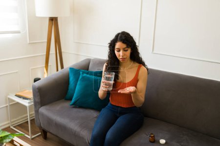 Téléchargez les photos : Malade jeune femme se sentant malade prendre des médicaments ou des vitamines avec un verre d'eau tout en se relaxant à la maison - en image libre de droit