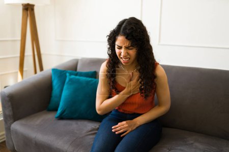 Foto de Mujer joven enferma que tiene dolor en el pecho o un ataque al corazón después de tener problemas cardíacos solos en casa - Imagen libre de derechos