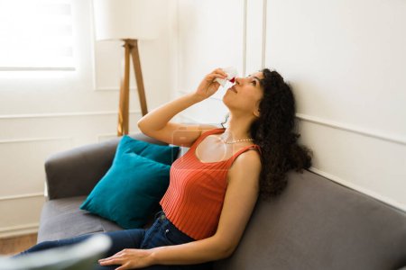 Foto de Mujer joven y cansada descansando en el sofá poniendo la cabeza hacia atrás con una hemorragia nasal y usando un pañuelo para sangrar la nariz - Imagen libre de derechos