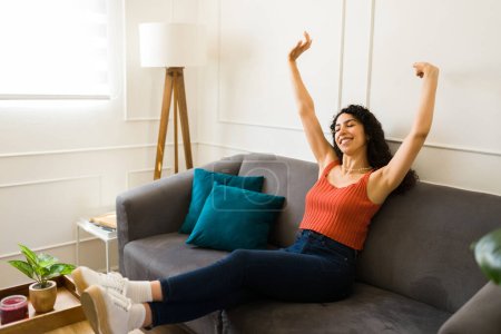 Foto de Relajada hermosa mujer latina levantando los brazos y relajándose mientras descansa en el sofá en su nueva casa de la sala de estar - Imagen libre de derechos