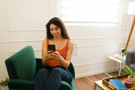 Téléchargez les photos : Femme hispanique joyeuse dans la vingtaine se détendre tout en se reposant dans le salon et textos ou en utilisant les médias sociaux sur le smartphone - en image libre de droit