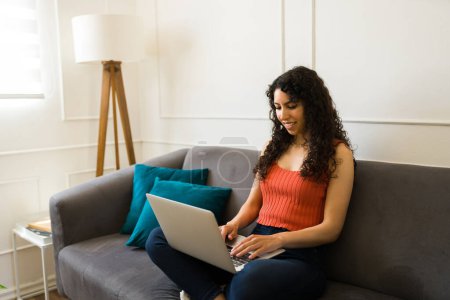 Photo for Cheerful beautiful young woman typing on the laptop while relaxing on the couch and going online - Royalty Free Image