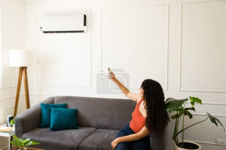 Téléchargez les photos : Latino jeune femme allumant le climatiseur dans son salon tout en se sentant chaud pendant l'été - en image libre de droit