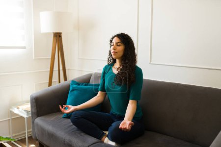 Foto de Mujer joven y relajada escuchando música de meditación y haciendo yoga o ejercicios de respiración en casa sentada en el sofá - Imagen libre de derechos