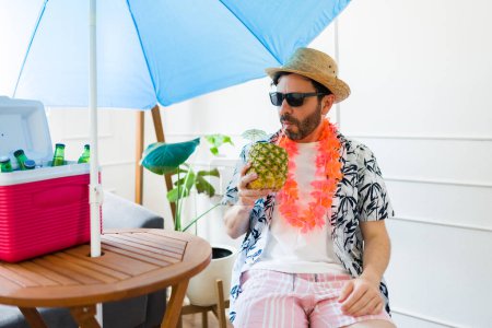 Téléchargez les photos : Homme attrayant avec des lunettes de soleil profiter de boire une boisson à l'ananas et se détendre à la maison en prétendant être à la plage d'été - en image libre de droit