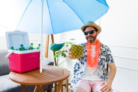 Téléchargez les photos : Gai hispanique homme disant acclamations boire un verre d'ananas et l'air heureux de son séjour d'été - en image libre de droit