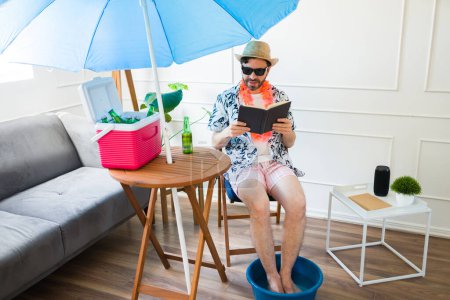 Foto de Sonriente hombre relajado leyendo un libro mientras se hace una pedicura en casa mientras disfruta de sus vacaciones de verano fingiendo estar en la playa - Imagen libre de derechos