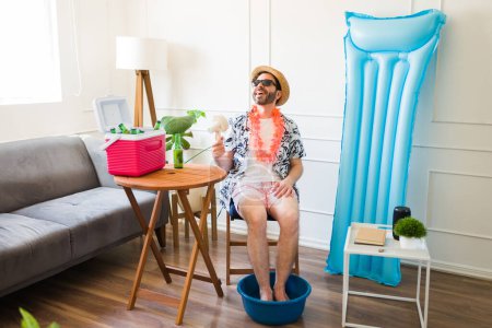 Entspannter glücklicher Mann lächelt bei einer Pediküre zu Hause, während er in den Sommerferien einen Ventilator mit Shorts und Sonnenbrille benutzt
