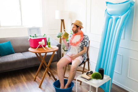 Photo for Caucasian mid-adult man drinking a pineapple drink and having a staycation in the living room looking relaxed with inflatables and a cooler - Royalty Free Image