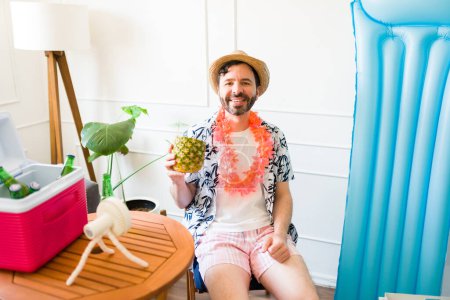 Téléchargez les photos : Joyeux bel homme mi-adulte avec une boisson à l'ananas souriant tout en profitant d'un séjour d'été relaxant à la maison - en image libre de droit