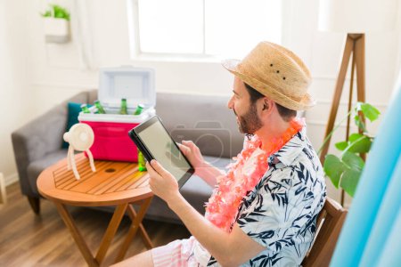 Téléchargez les photos : Joyeux homme mi-adulte avec un chapeau d'été vu de derrière souriant regardant une vidéo et utilisant une tablette pendant ses vacances à la maison - en image libre de droit