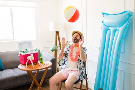 Foto de Hombre caucásico divertido divirtiéndose jugando con una pelota de playa en la sala de estar mientras finge estar en unas vacaciones de verano en casa - Imagen libre de derechos