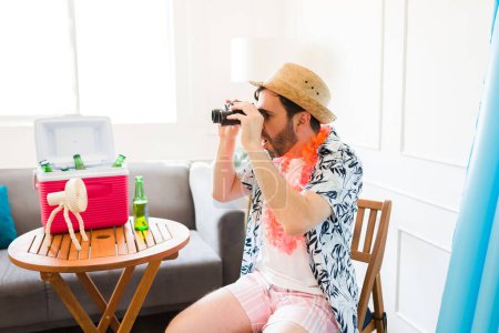 Foto de Perfil de un hombre caucásico que luce sorprendido usando prismáticos en la sala de estar durante una estancia de verano con bebidas y un refrigerador - Imagen libre de derechos