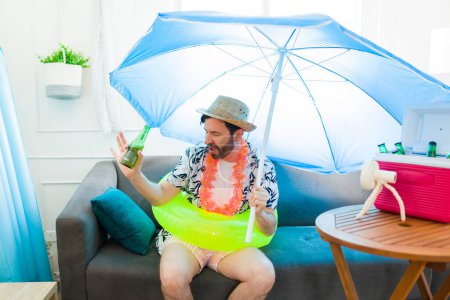 Foto de Divertido hombre adulto medio bailando usando un inflable y sombrilla mientras bebe una cerveza en la sala de estar fingiendo estar en vacaciones en la playa - Imagen libre de derechos