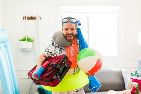 Téléchargez les photos : Latin excité homme regardant surpris et heureux tout en portant des lunettes de plongée, ballon de plage et valise pour aller à ses vacances d'été - en image libre de droit