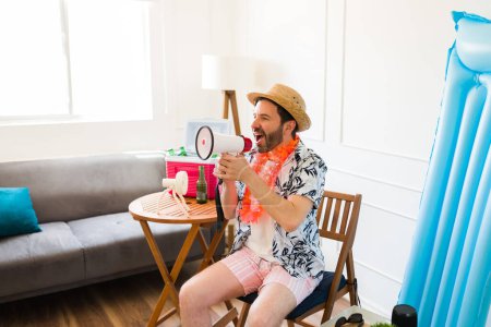 Foto de Atractivo hombre feliz usando un megáfono gritando en la sala de estar mientras que quiere viajar a la playa de vacaciones - Imagen libre de derechos