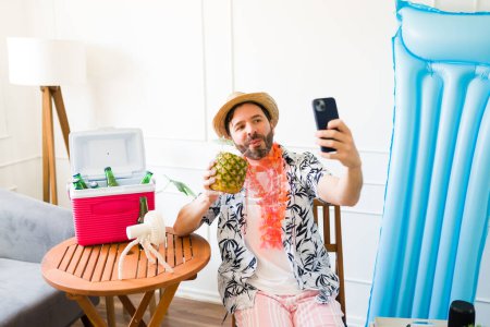 Photo for Funny mid-adult man with a hat taking a selfie with his phone while having a pineapple drink in the living room during staycations - Royalty Free Image